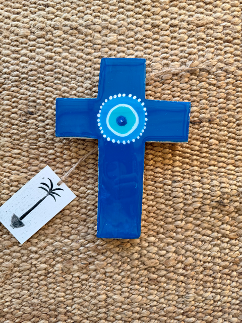 A blue painted cross ornament with a hand-painted evil eye design in the center, resting on a beige woven jute rug with a small white tag attached.
