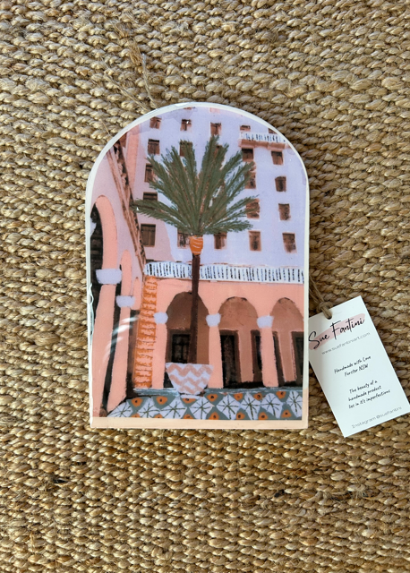 A handcrafted tile with a depiction of a building and a palm tree in colors pink, beige, and green, with a metal hook on the back.