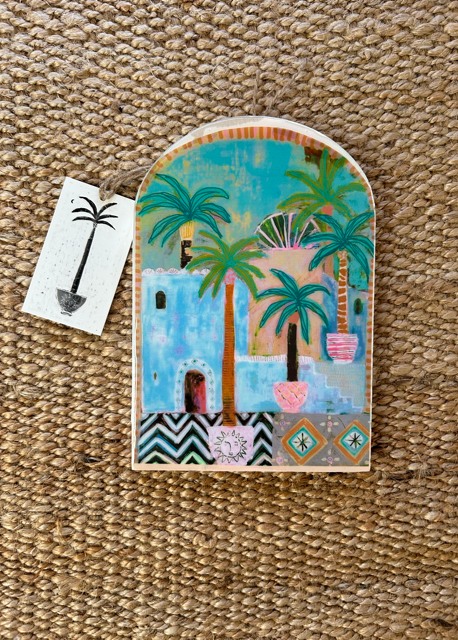 A handcrafted ceramic tile with a painting of green palm trees and a blue building on a beige background, marked with 'Potted Palms' and a size indication of '22 x 15cm'. A metal hook is attached to the back for hanging.