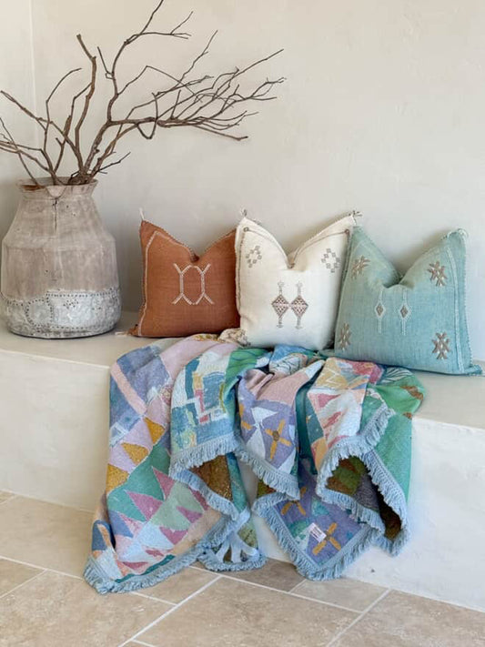Colorful quilt and pillows on a bench with a vase and branches in the background.
