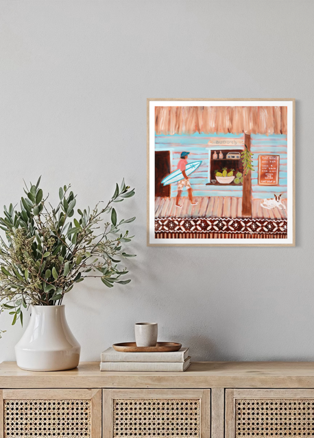 A framed art print depicting a beach house scene with a person carrying a surfboard, displayed on a light gray wall above a wooden cabinet with a vase of greenery and a small cup.