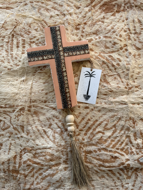 A plaster cross ornament with peach-colored sides, black patterned borders, a tassel, and a small hanging tag with a palm tree illustration, displayed on a textured beige fabric background.