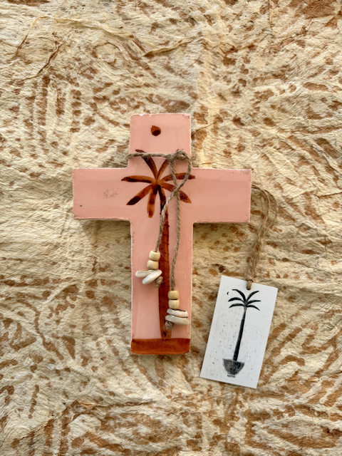 A handmade plaster cross ornament in a dusty pink color, decorated with a brown palm tree, small wooden beads, and a hanging string. A small white tag with a black palm tree illustration is attached.