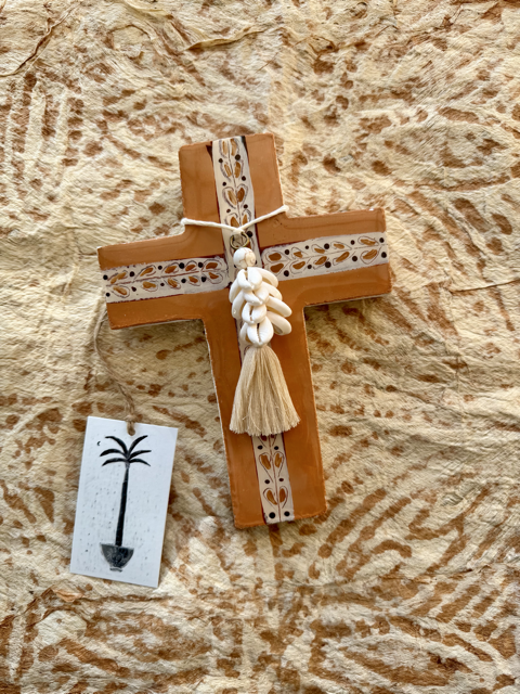 A plaster cross ornament with carved floral details, a white shell centerpiece, and a beige tassel, displayed on a textured beige surface next to a small white tag with a black palm tree illustration.