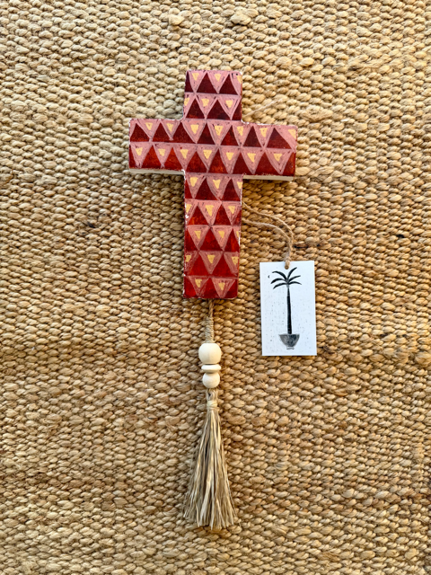 A painted plaster cross ornament with a geometric triangular pattern in reddish-brown and beige, featuring a natural wood tassel and a small white tag with a palm tree illustration, displayed on a textured beige woven rug.