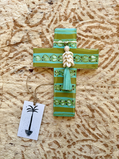 A handmade cross-shaped ornament made of layered green and turquoise striped material, decorated with a small white shell bead and tassel, shown on a textured tan background with a small hanging tag.