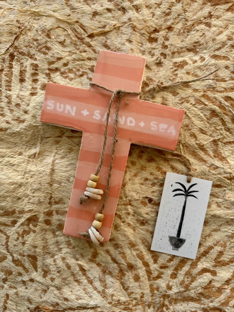 A handmade plaster cross ornament painted in a coral pink and orange striped pattern, featuring the words 'SUN + SAND + SEA' in white. It is decorated with small white and beige seashells and a piece of natural twine.
