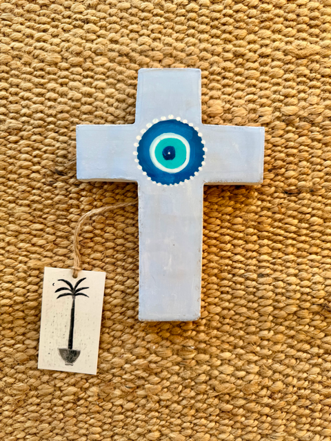 A hand-painted plaster cross in a light blue-gray finish, decorated with a blue and white evil eye design in the center. It is displayed on a textured beige woven rug with a small wooden tag hanging from its side.
