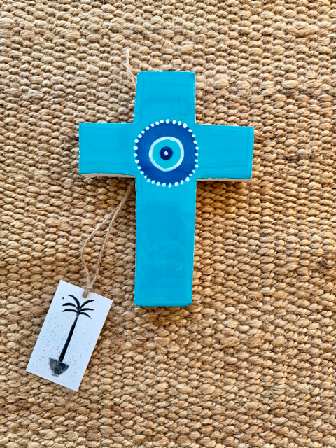A painted plaster cross ornament in light blue with a circular blue and white evil eye design in the center, hanging on a woven beige jute rug with a small white tag below it.