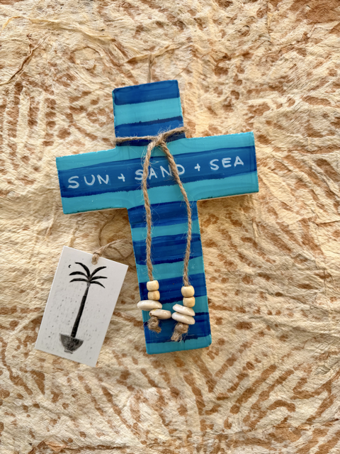 A painted plaster cross ornament in shades of blue and teal with the words 'SUN + SAND + SEA' on the front, tied with twine and decorated with small white and natural beads. A small tag with a palm tree illustration hangs from the cross, placed on a textured tan surface.
