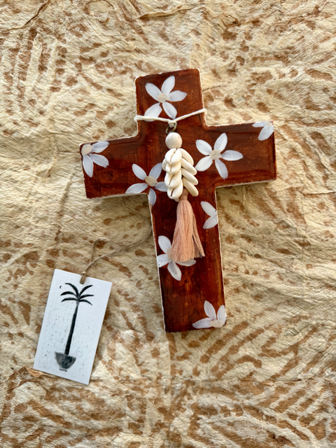A plaster cross ornament with white floral inlays, a white shell bead garland, and a pink tassel, displayed on a textured beige paper background with a small white tag.