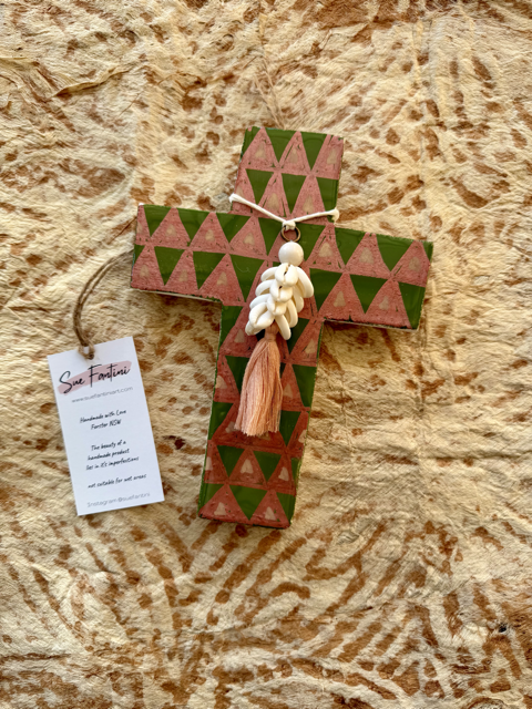 A handcrafted plaster cross ornament with a green and pink geometric triangle pattern, decorated with a white shell bead and a pink tassel