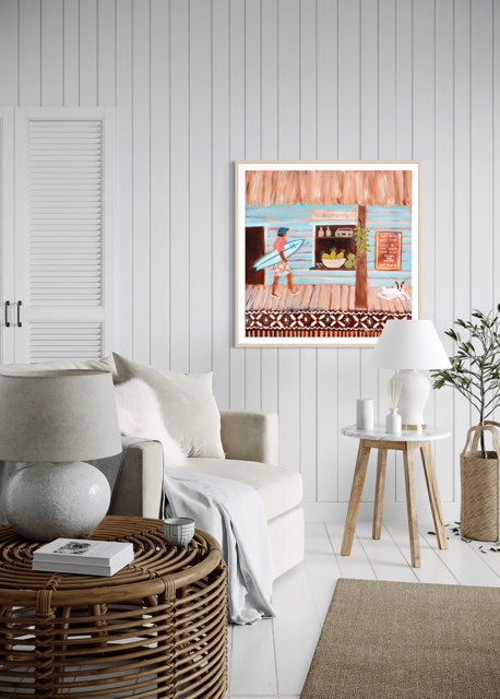 A framed art print depicting a colorful beach hut scene with a surfer, hung on a white paneled wall in a modern, sunlit living room with a sofa, side table, and plants.