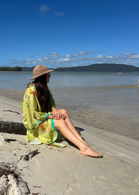 Woman in a green kimono and hat sitting on a beach with a scenic background
