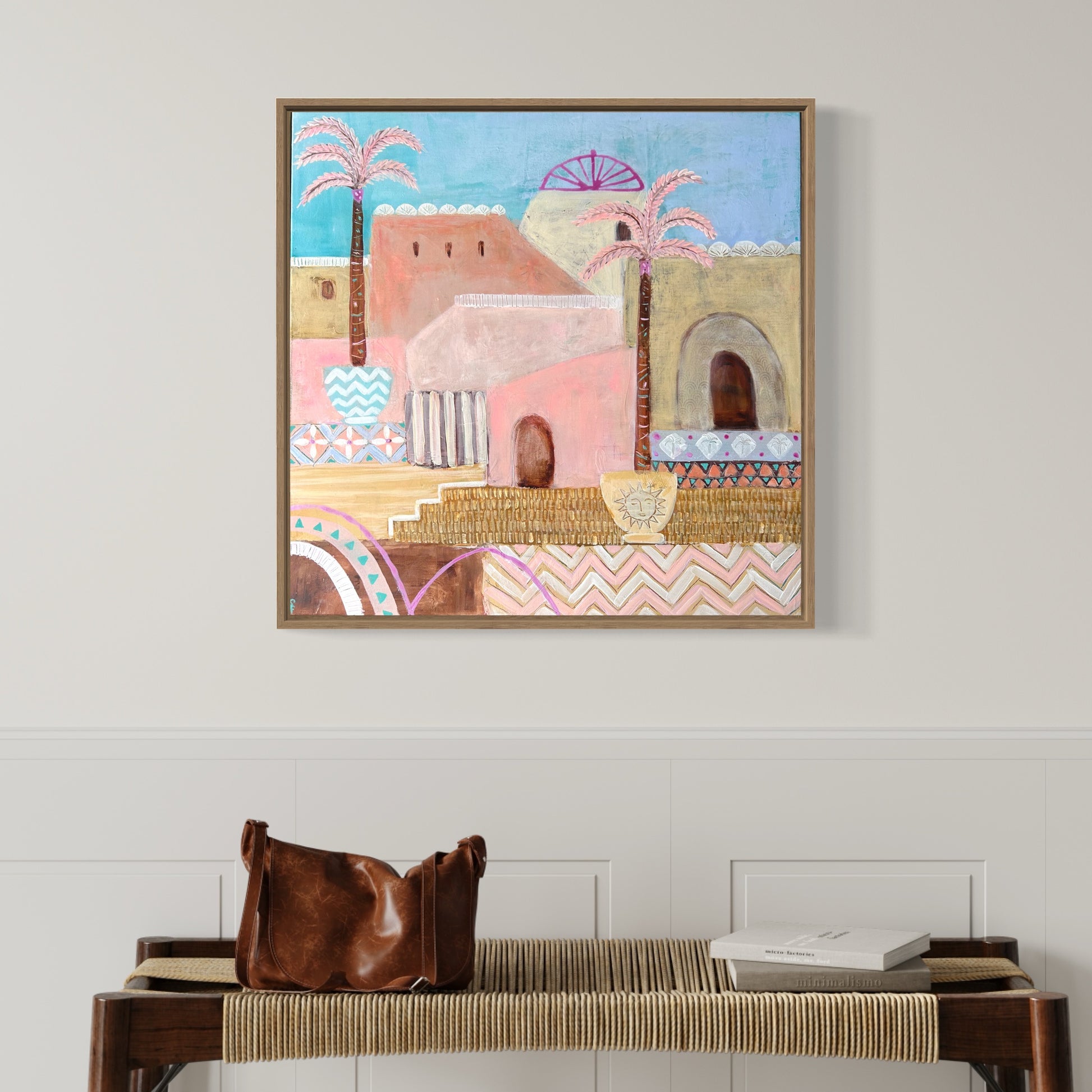 Original artwork in pinks and yellows of Moroccan median with palm trees, framed on wall in front of side table with leather handbag